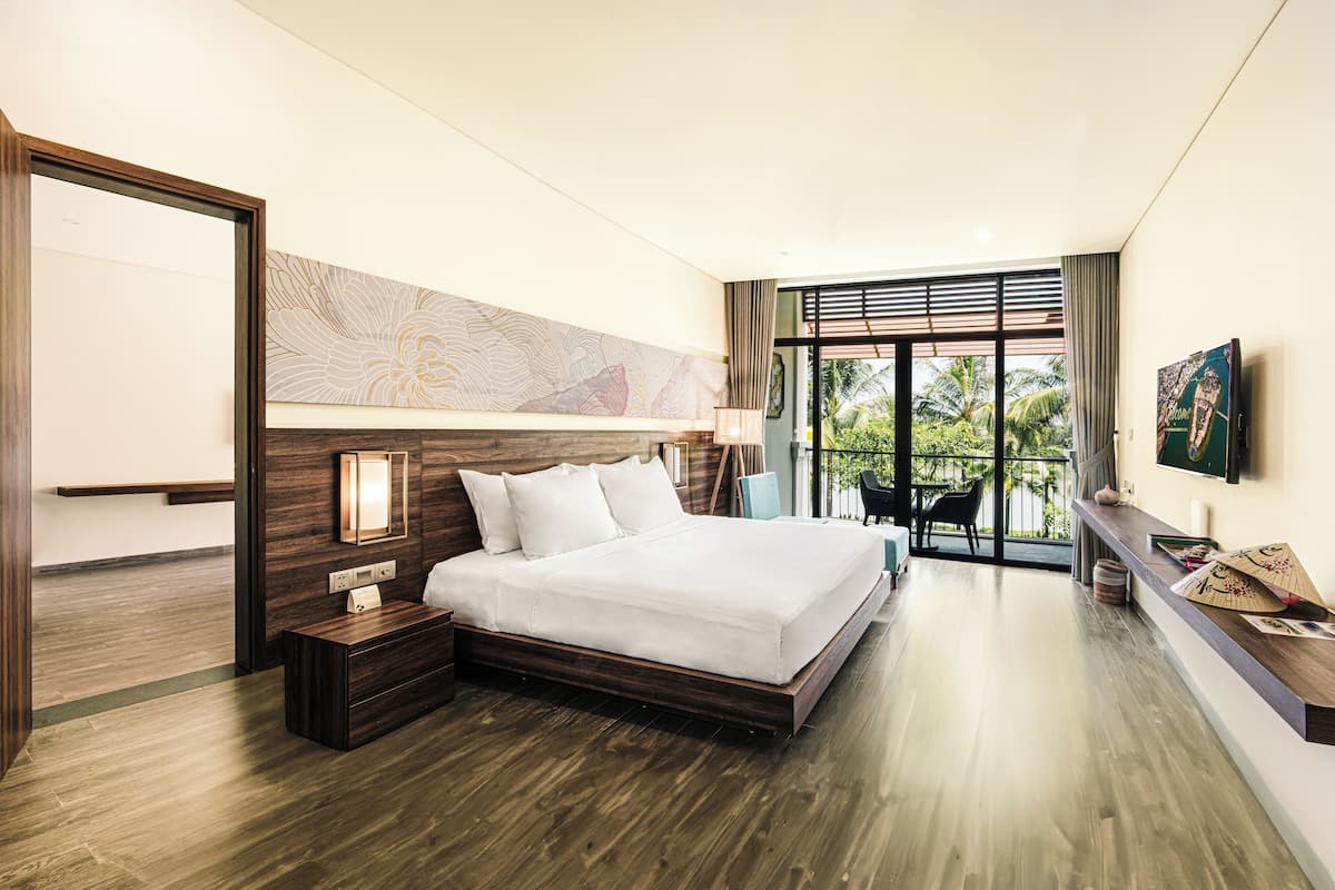 A modern hotel room with a large bed, wooden flooring, a view of the outdoors, and a TV mounted on the wall.