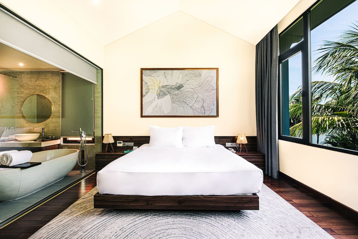 A modern hotel room with a large bed, a built-in bathtub, a large window with a view of palm trees, and a wall-mounted artwork.