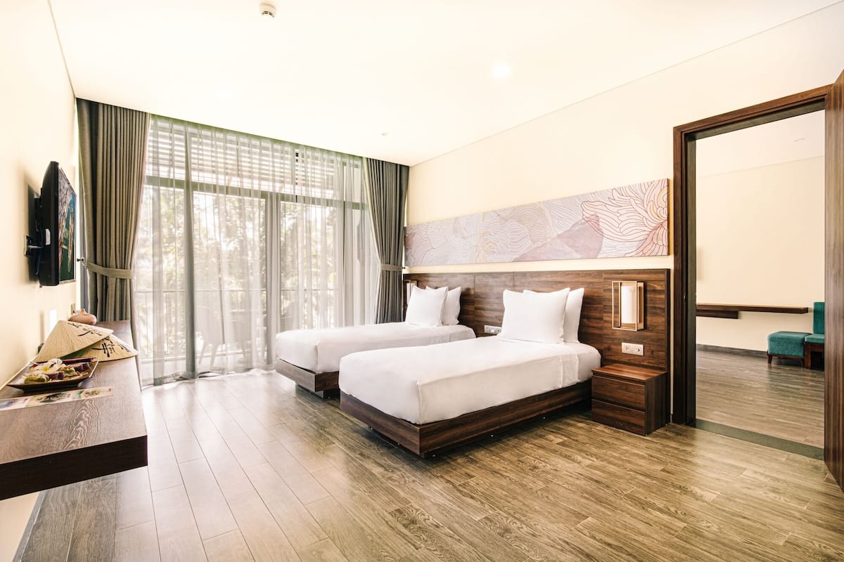 A hotel room with two beds, a wooden floor, a large window with curtains, a flat-screen TV, and a decorative wall piece.