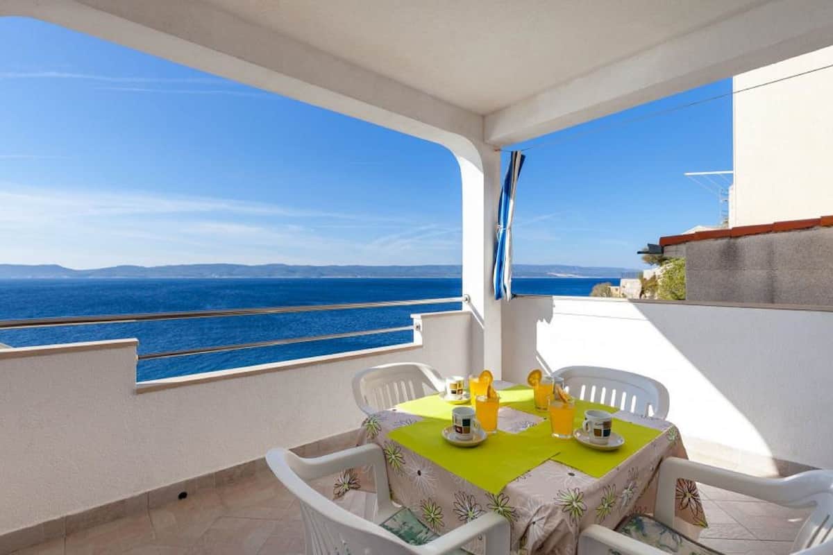 A balcony with a table set for two, overlooking a clear blue sea and distant land.