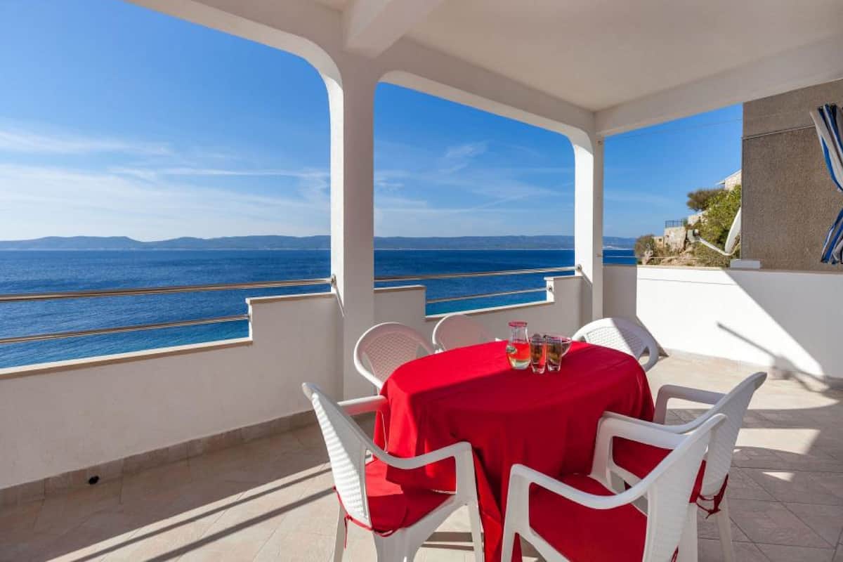 A covered outdoor seating area with white chairs and a table set with a red tablecloth, overlooking a coastal view.