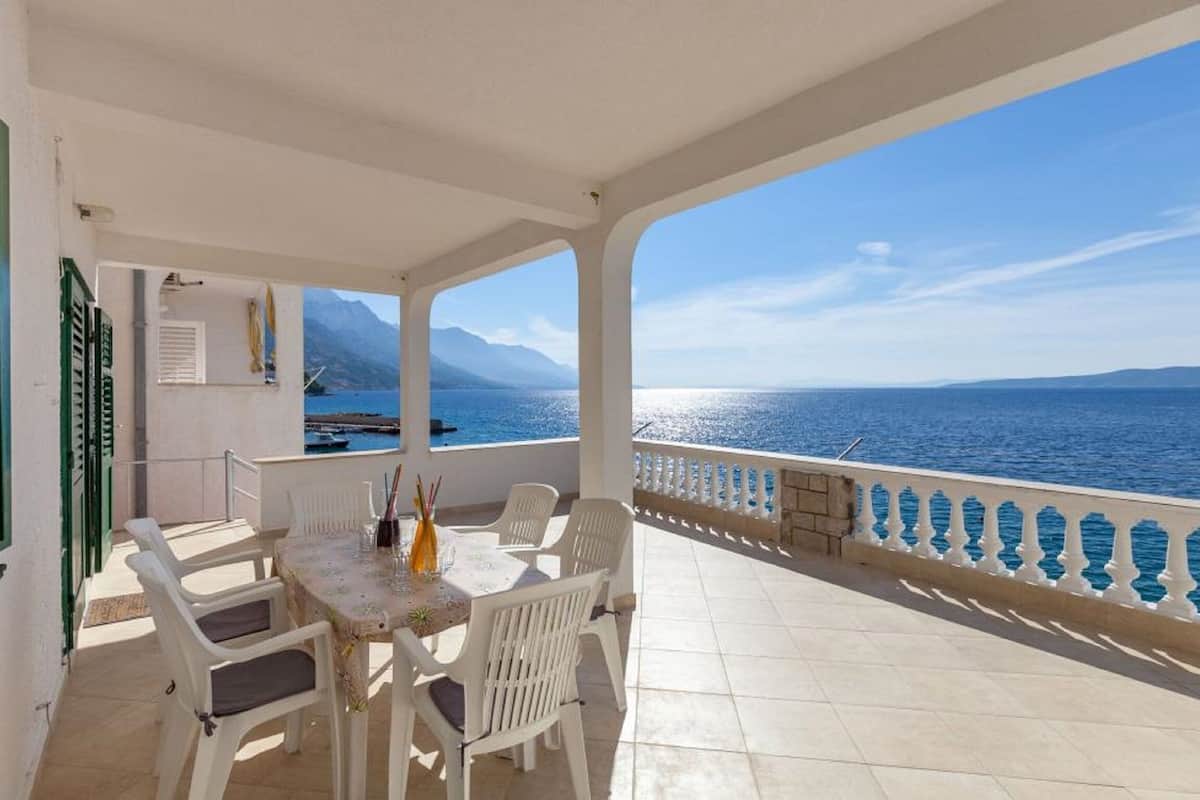 A covered outdoor area with a table and chairs, a view of the sea, and a balcony with a railing.
