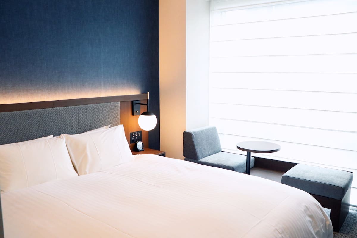 Hotellihuone, jossa on suuri sänky, yöpöytä lampun kanssa, pieni pyöreä pöytä ja tuoli.