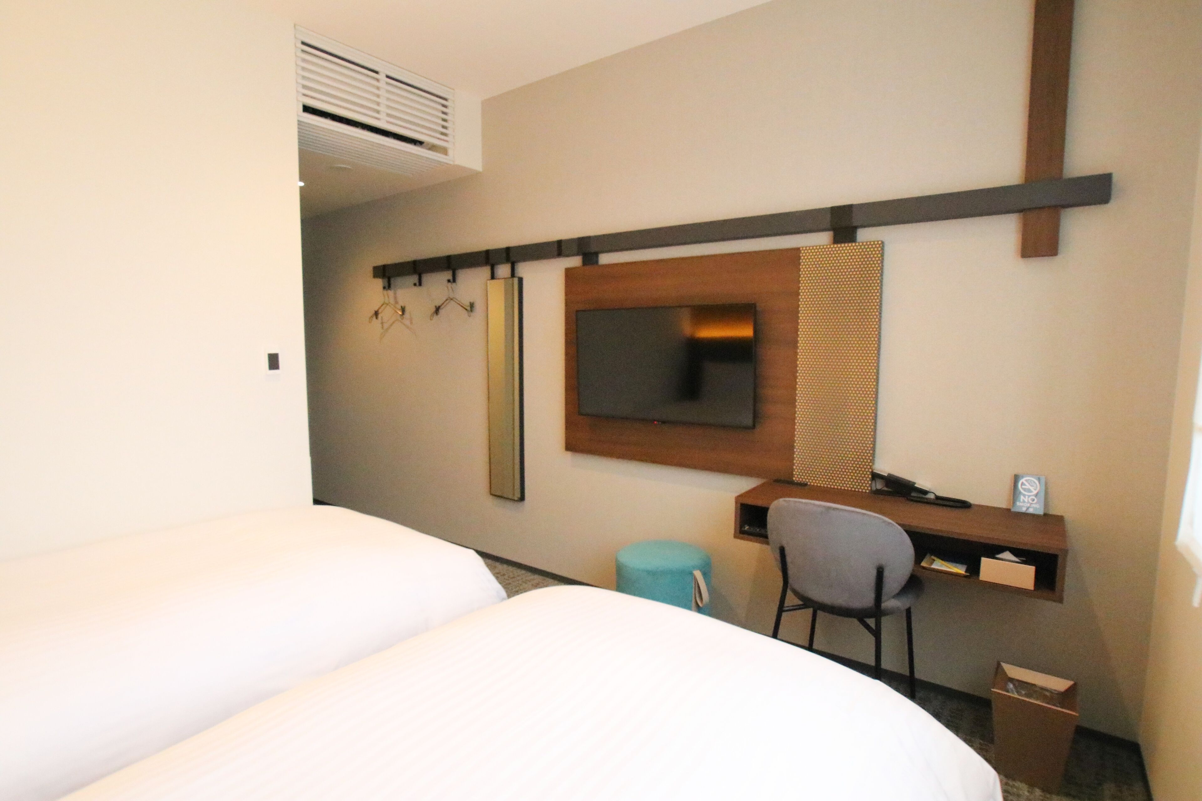 A hotel room with two beds, a desk with a chair, a television, and a small stool.