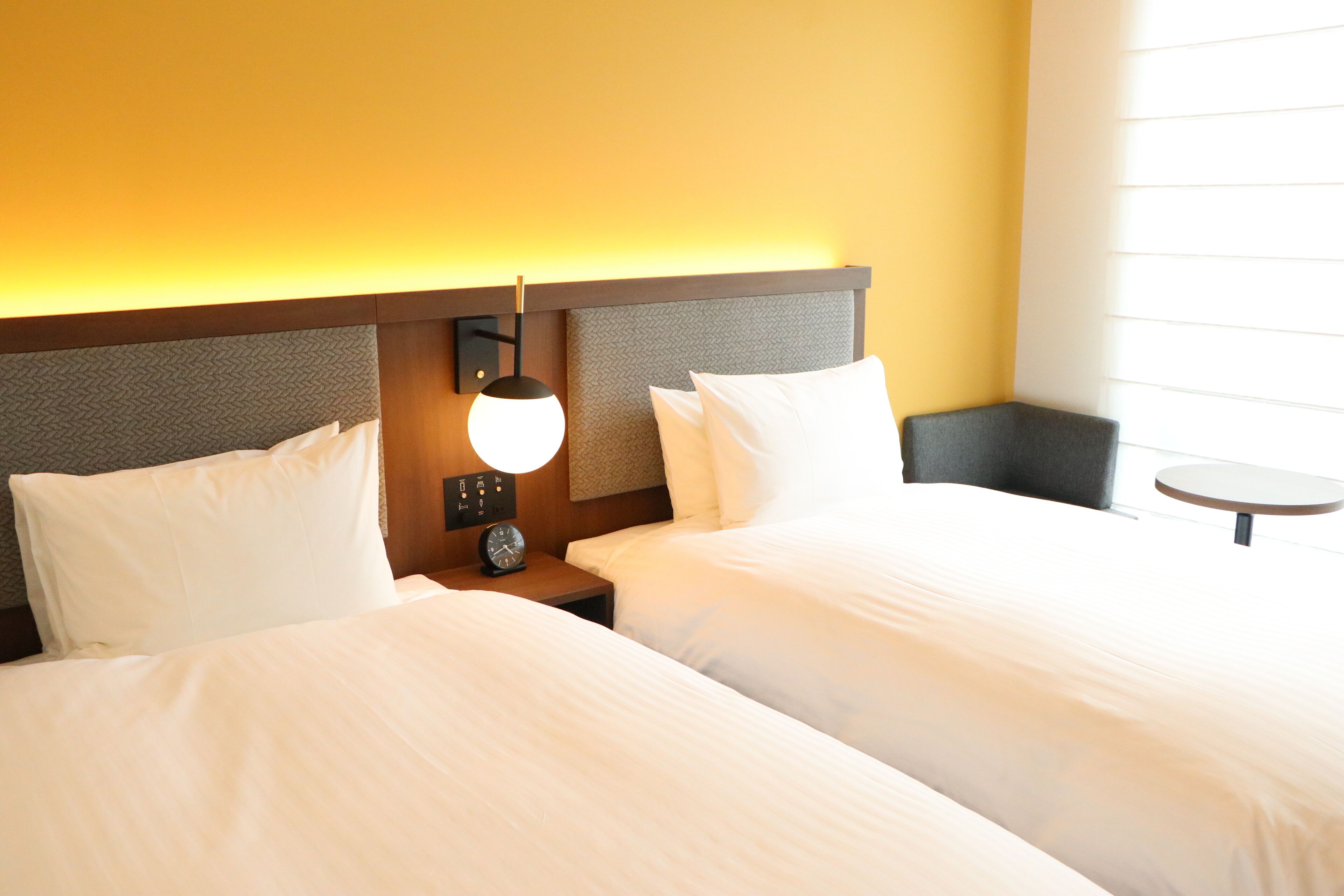 Habitación de hotel con dos camas, una mesita de noche con lámpara, un reloj y una pequeña mesa redonda.