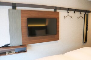 Ein modernes Hotelzimmer mit einem an der Wand befestigten Flachbildfernseher, einer Wandverkleidung aus Holz, einer kleinen Ablage mit Fernbedienung und mehreren Wandhaken.