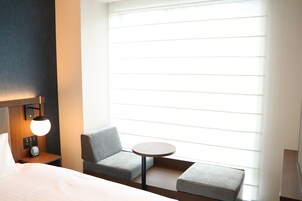 A hotel room with a bed, a small table, two armchairs, a nightstand with a lamp, and a large window with blinds.