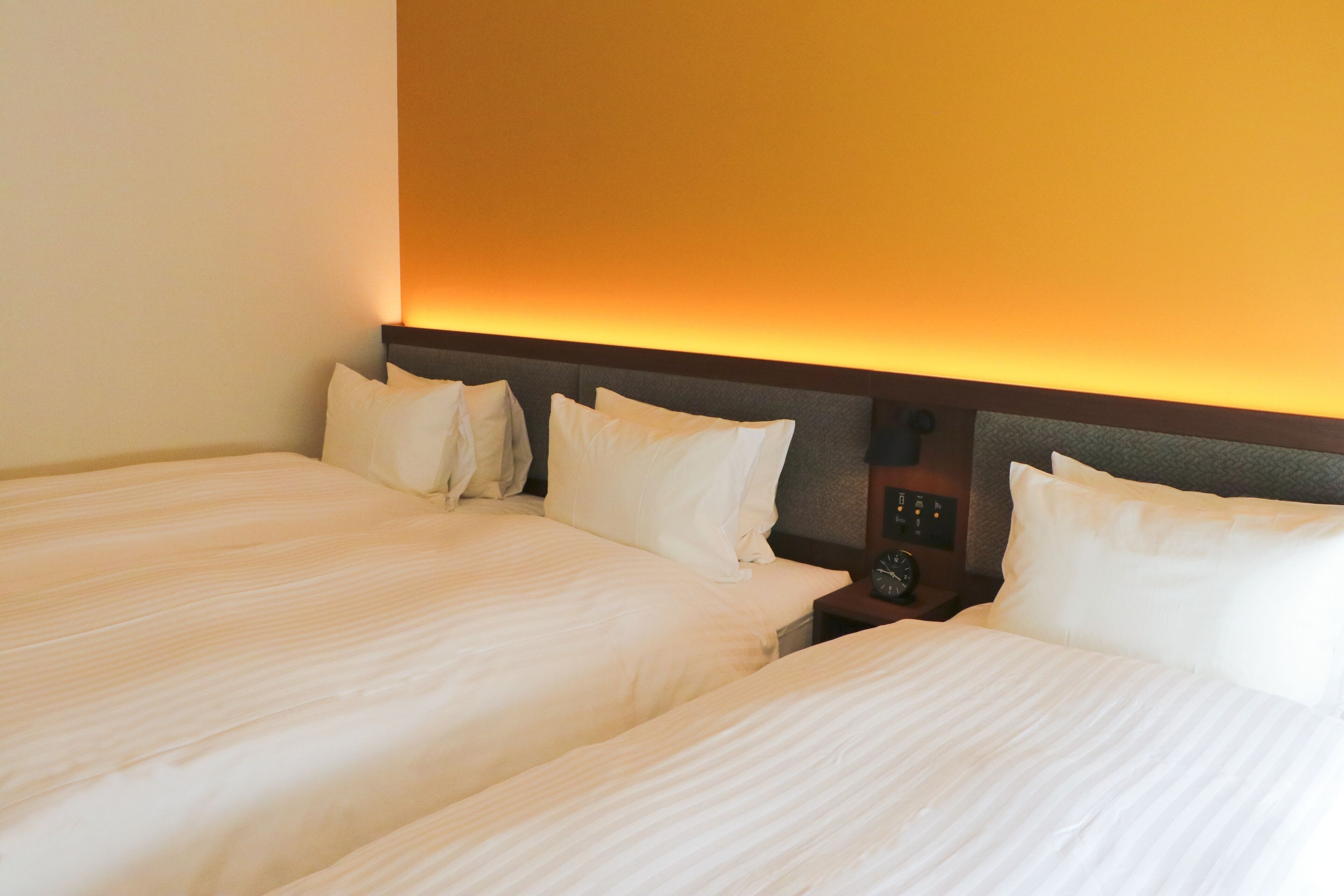 Habitación de hotel con dos camas, una mesita de noche con un reloj y un panel de control en la pared.