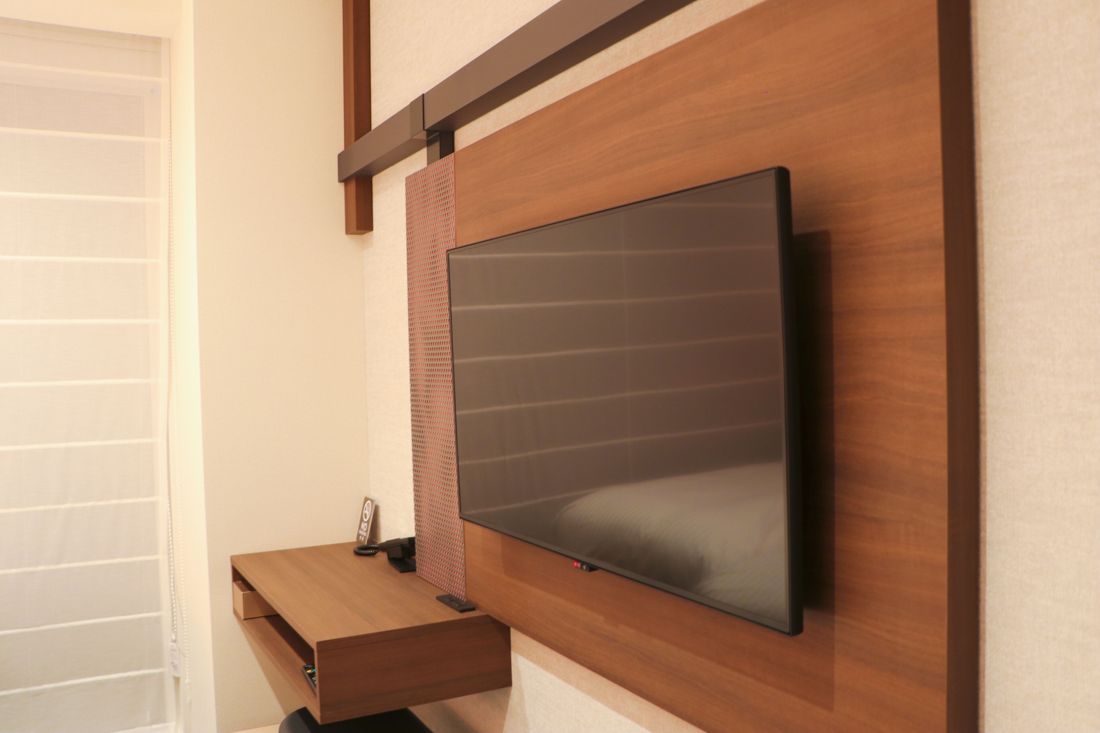 A modern room with a mounted flat-screen TV on a wooden wall, a wooden desk with a drawer, and a window with blinds.