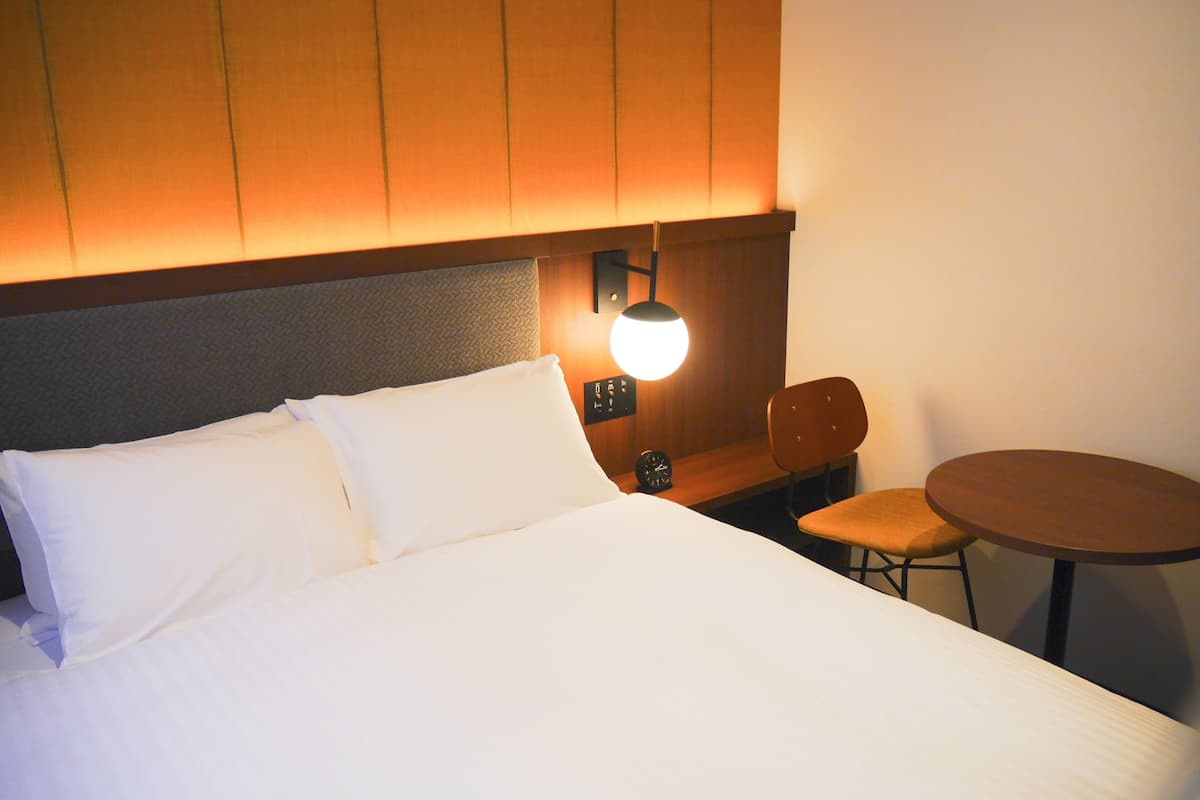 Hotellihuone, jossa on sänky, yöpöytä, jossa on kello, pieni pöytä ja tuoli.