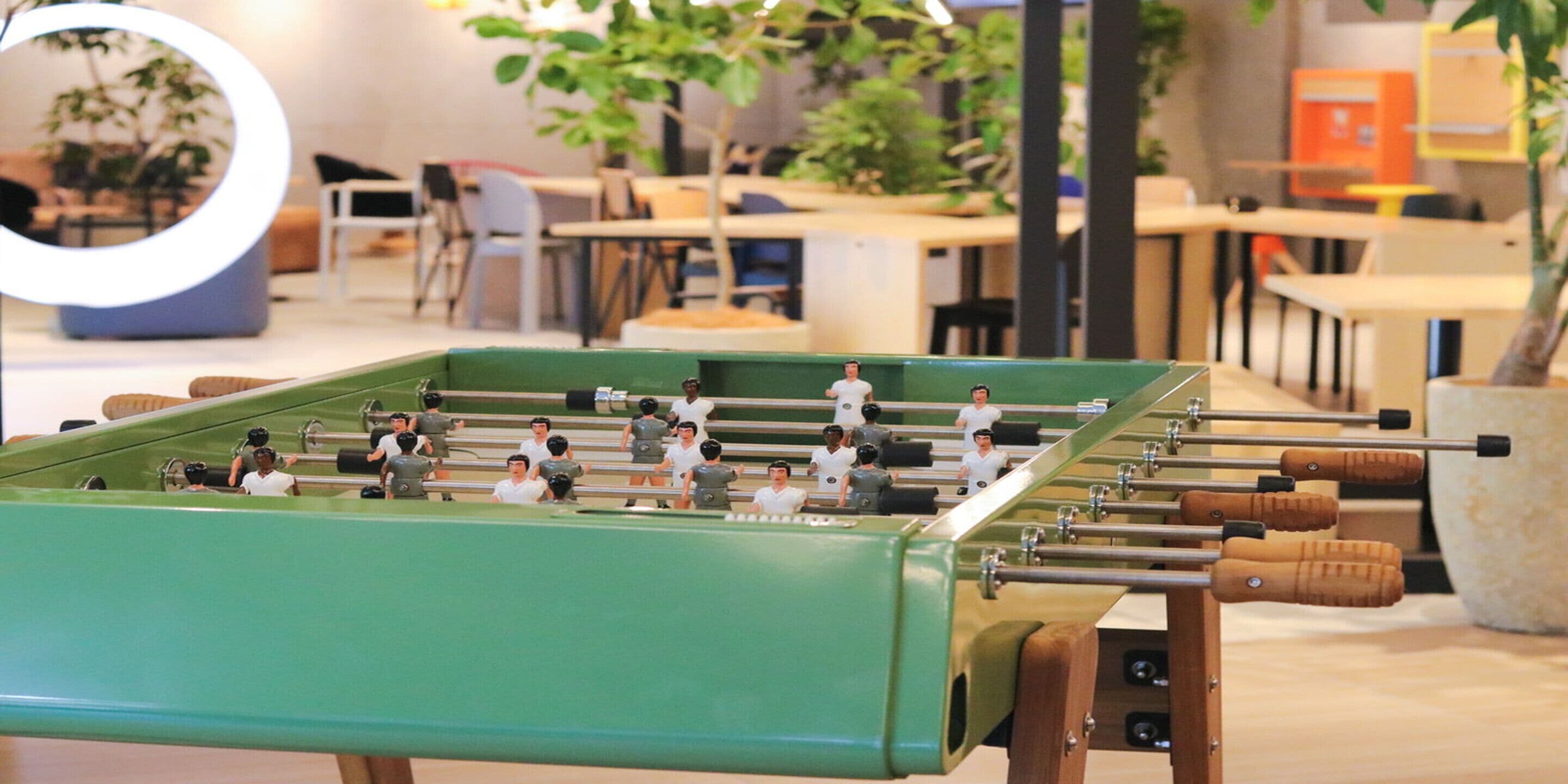 A foosball table in a room with tables and chairs, plants, and a circular light fixture.