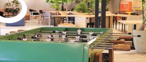 A foosball table in a room with tables and chairs, plants, and a circular light fixture.