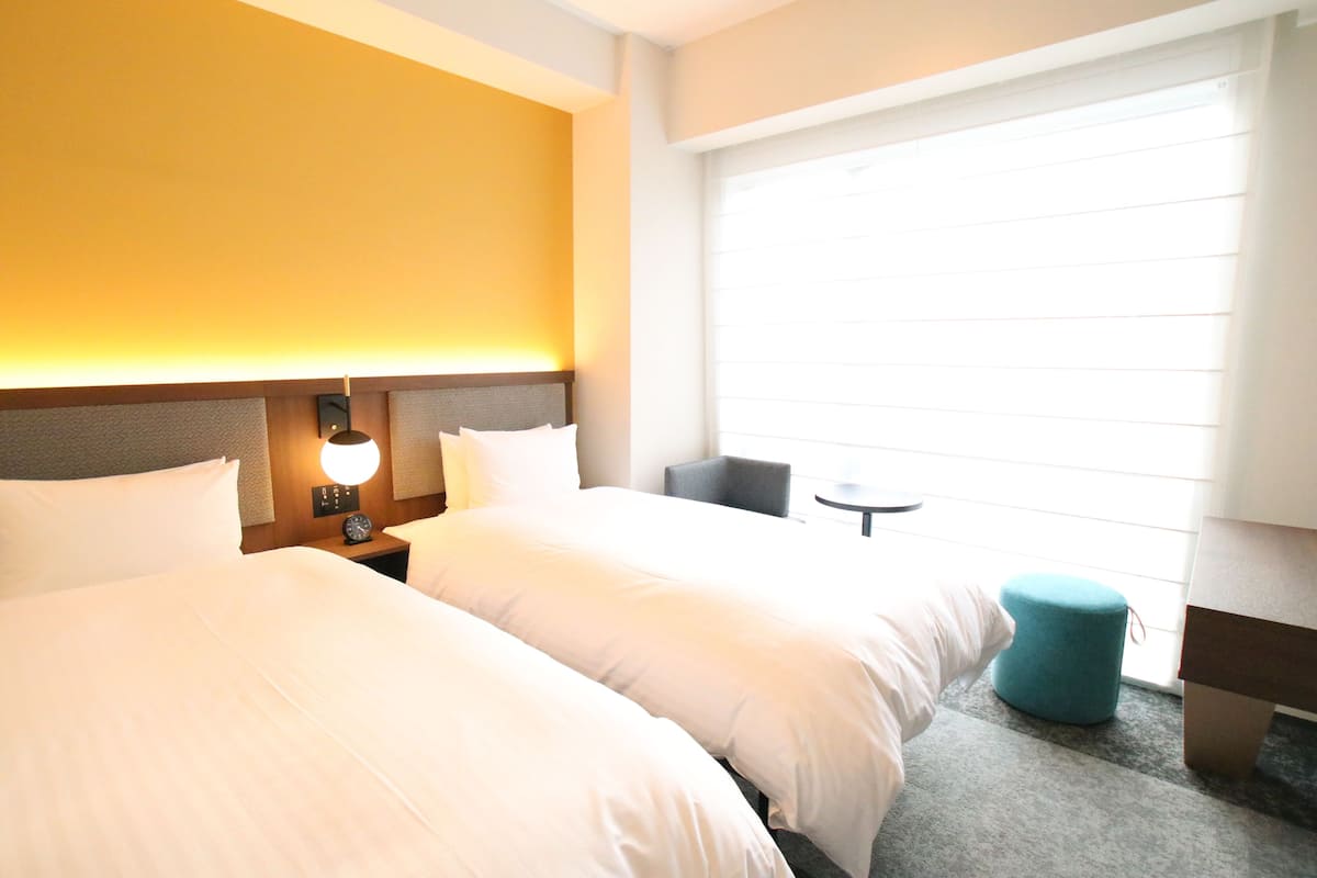 Hotellihuone, jossa on kaksi sänkyä, yöpöytä lampun kanssa, pieni pyöreä pöytä ja ikkuna, jossa on sälekaihtimet.
