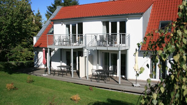 A two-story building with a red-tiled roof, a balcony with railings, and a covered outdoor seating area with tables and chairs.