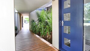 Um deck de madeira leva a uma porta azul com painéis de vidro, em meio à vegetação.