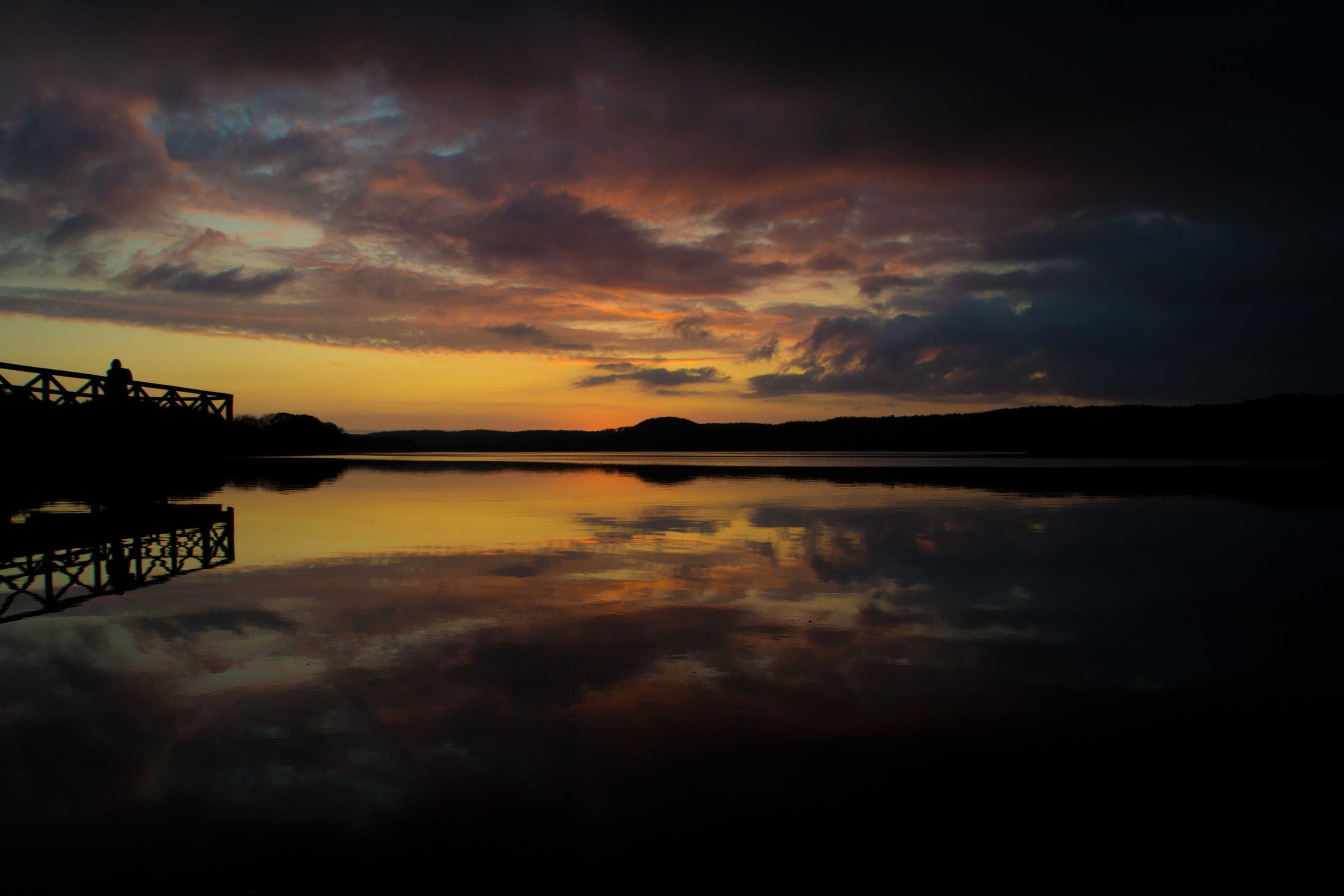 A sunset over a calm body of water with a dock and distant hills.