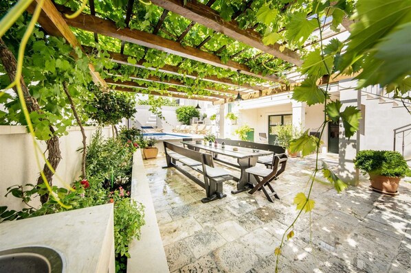 A covered outdoor seating area with a pool, surrounded by greenery and potted plants.