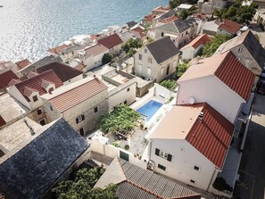 A coastal residential area with terracotta-roofed buildings, a central swimming pool, and a view of the sea.