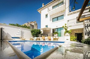 A swimming pool with lounge chairs, a white building with balconies, and a clear blue sky.