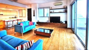 Una sala moderna con un sofá azul, una mesa de centro y un televisor. Cuenta con una zona de cocina con taburetes de bar y un ventanal grande.