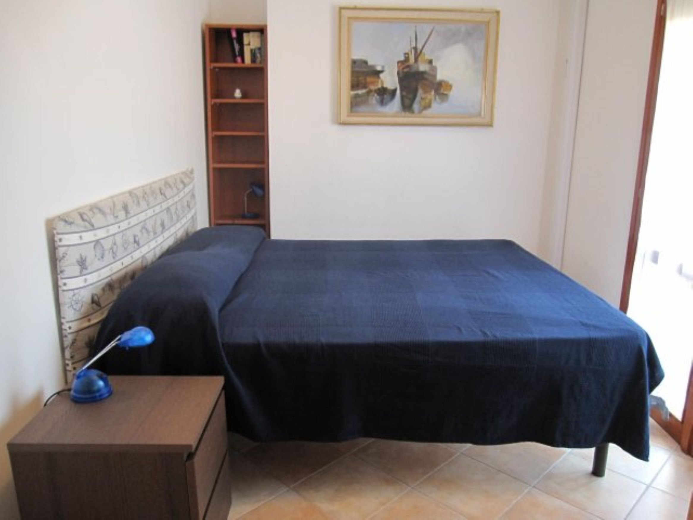 A bedroom with a bed, a nightstand, a lamp, a bookshelf, and a painting on the wall.