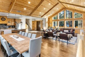 A spacious living area with a large wooden dining table, comfortable seating, and a view through large windows.