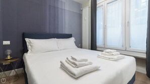 A neatly made bed with white linens and folded towels, a bedside table with a lamp, and a window with curtains. - Italianway - Garibaldi 104 C (Milan)