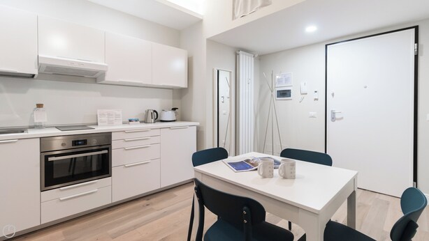 A modern kitchen with white cabinets, a built-in oven, and a dining area with a white table and chairs. - Italianway - Garibaldi 104 C (Milan)