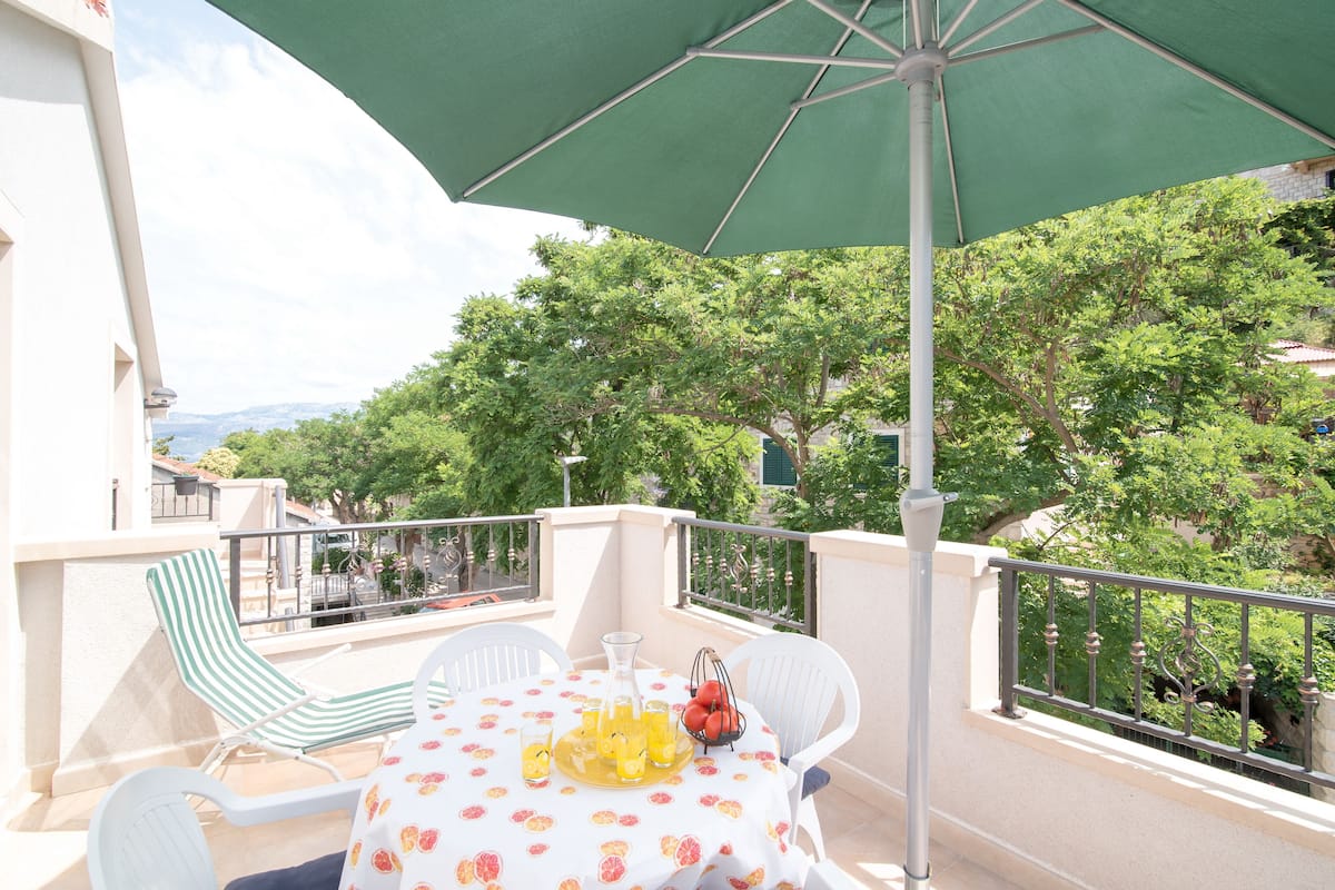 A balcony with a table set for two, a green umbrella, and a view of trees and buildings.