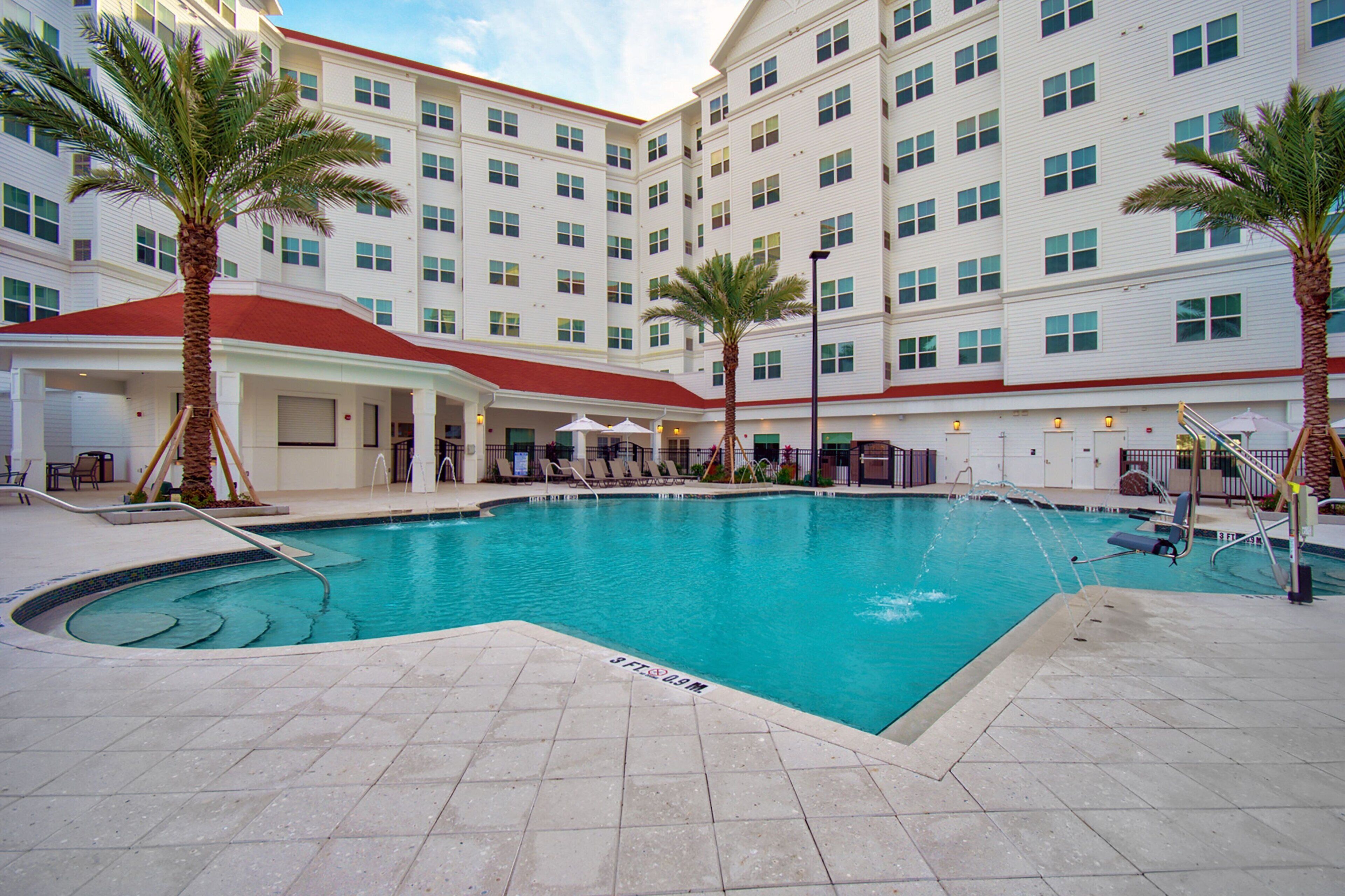 A hotel with a swimming pool, palm trees, and a covered area with lounge chairs.