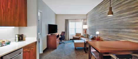 A modern hotel room with a kitchen area, a dining table, and a living area with a sofa and a TV.