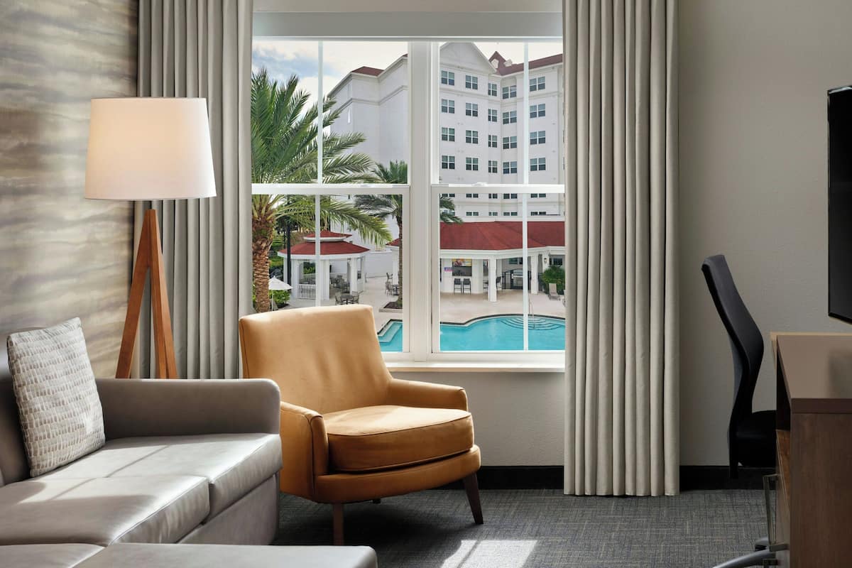 Habitación de hotel con sofá, sillón y una ventana grande con vistas a una piscina y edificios del hotel.
