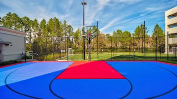 Una cancha de baloncesto al aire libre con superficie roja y azul, rodeada por una valla y árboles.