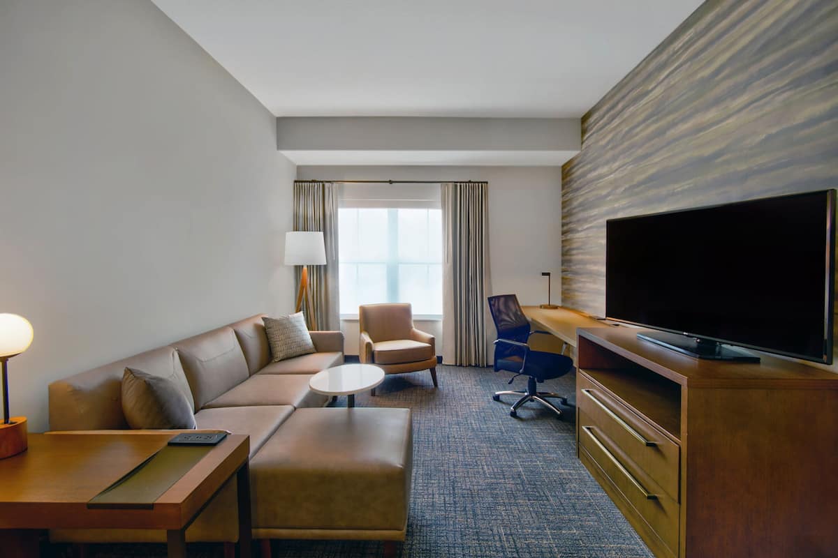 Una habitación de hotel con sofá, sillón, mesa de centro, televisor y escritorio con lámpara.