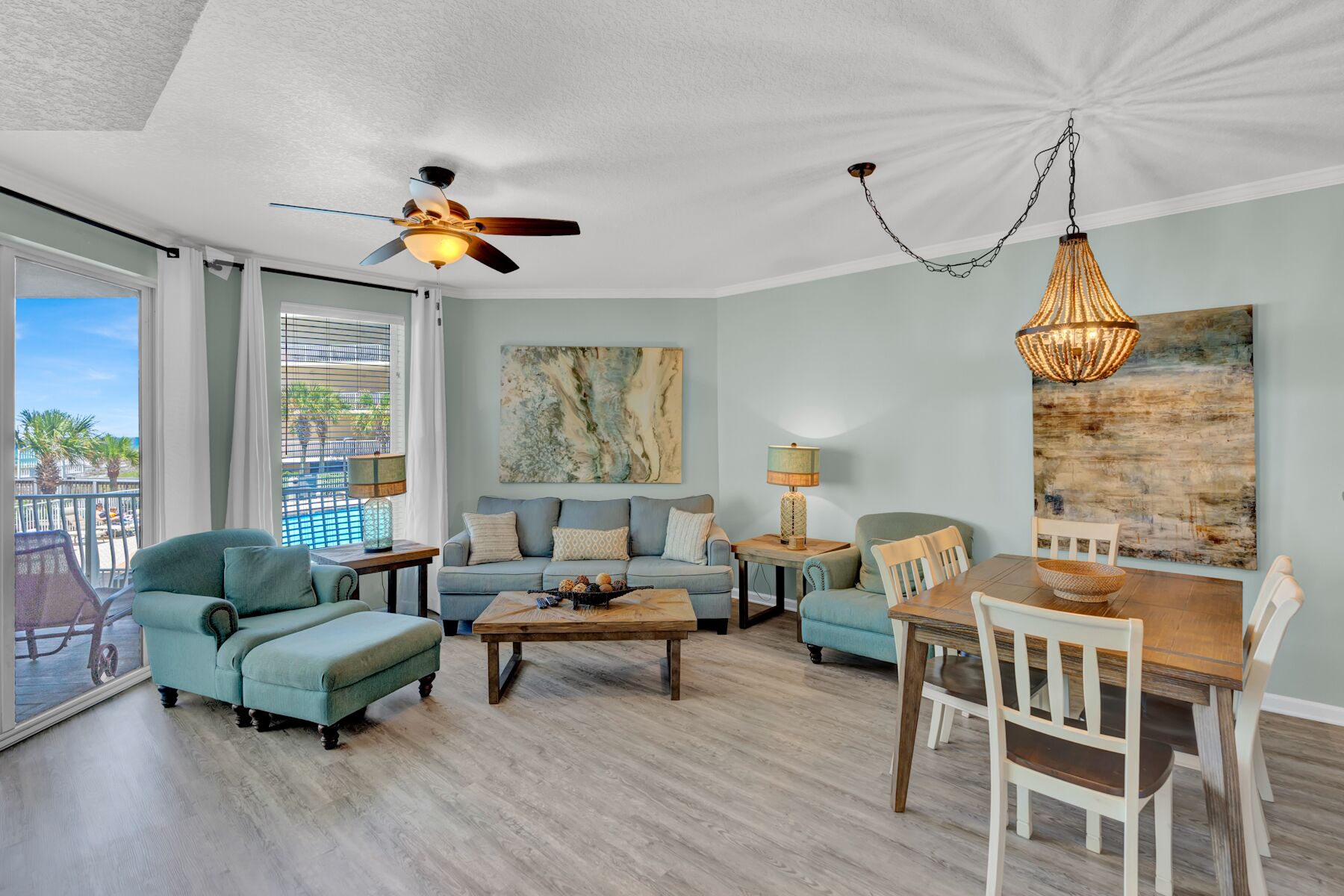 A spacious living room with a balcony, a ceiling fan, a dining table with chairs, and a large abstract painting on the wall.