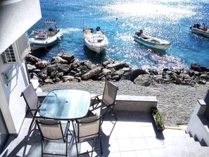 A terrace with a table and chairs overlooking a rocky shoreline with boats in the water.