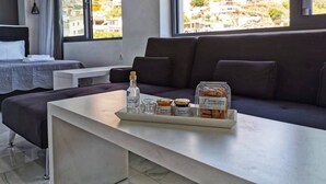 Habitación de hotel moderna con un amplio ventanal que ofrece vistas a un paisaje montañoso. Cuenta con una cama, un sofá y una mesita con una botella, vasos y un recipiente con aperitivos.