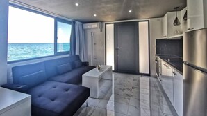 Un interior moderno con sofá, una pequeña mesa, una zona de cocina con refrigerador y vistas al mar.