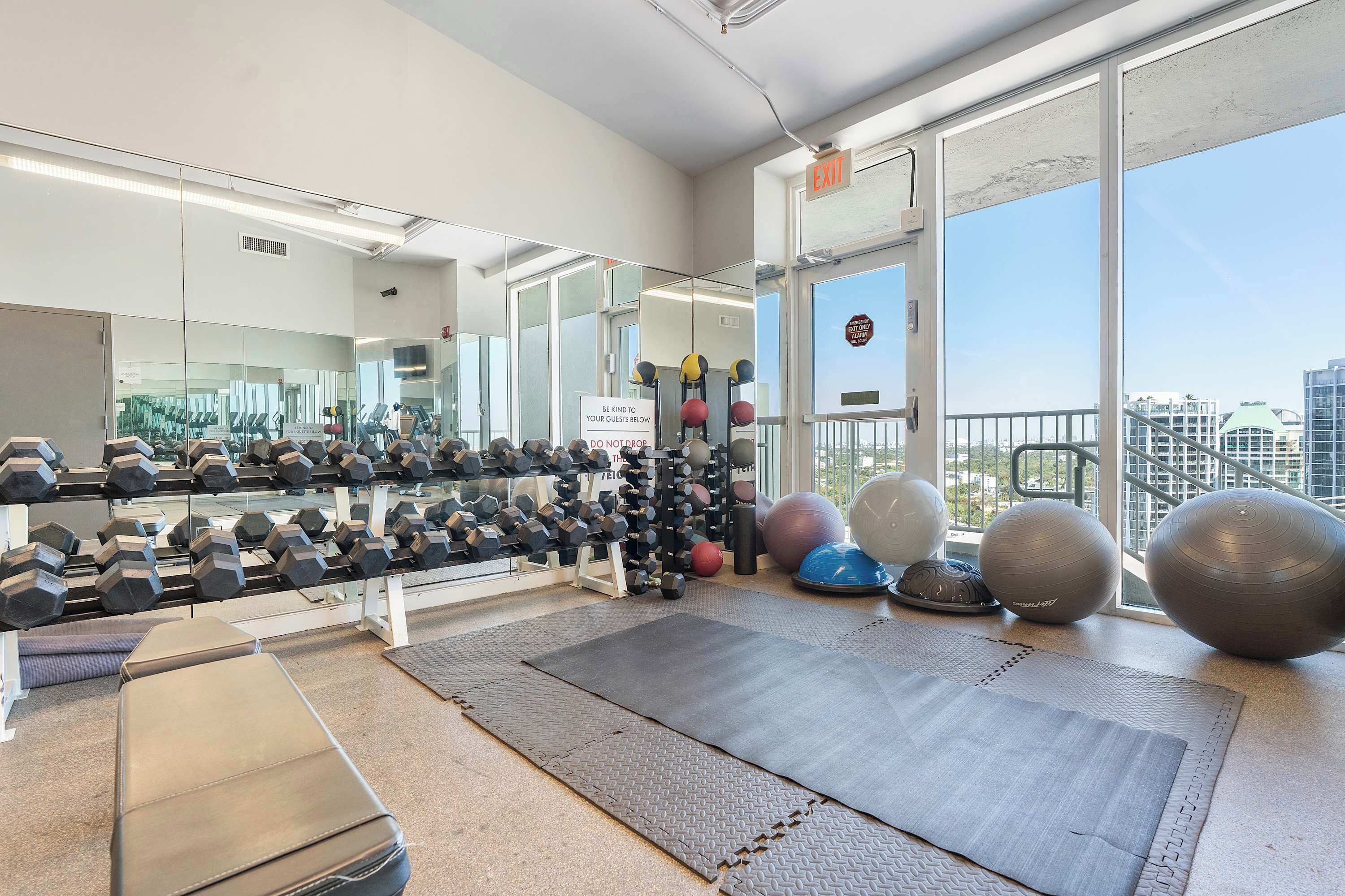 Un gimnasio con equipo de ejercicio, un espejo y grandes ventanales con vistas a la ciudad.