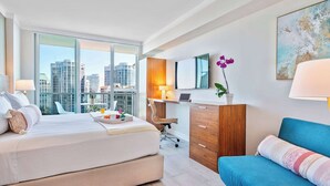 A modern hotel room with a large bed, a desk, a chair, a blue sofa, and a city view through large windows. - Panoramic Bay View Suite with Private Balcony (Miami)
