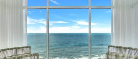 A room with a large window overlooking the ocean, two wicker chairs, and a stone sculpture.