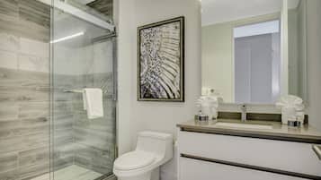 A modern bathroom with a glass shower enclosure, a toilet, a sink with a mirror, and a framed abstract artwork on the wall.