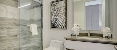 A modern bathroom with a glass shower enclosure, a toilet, a sink with a mirror, and a framed abstract artwork on the wall.
