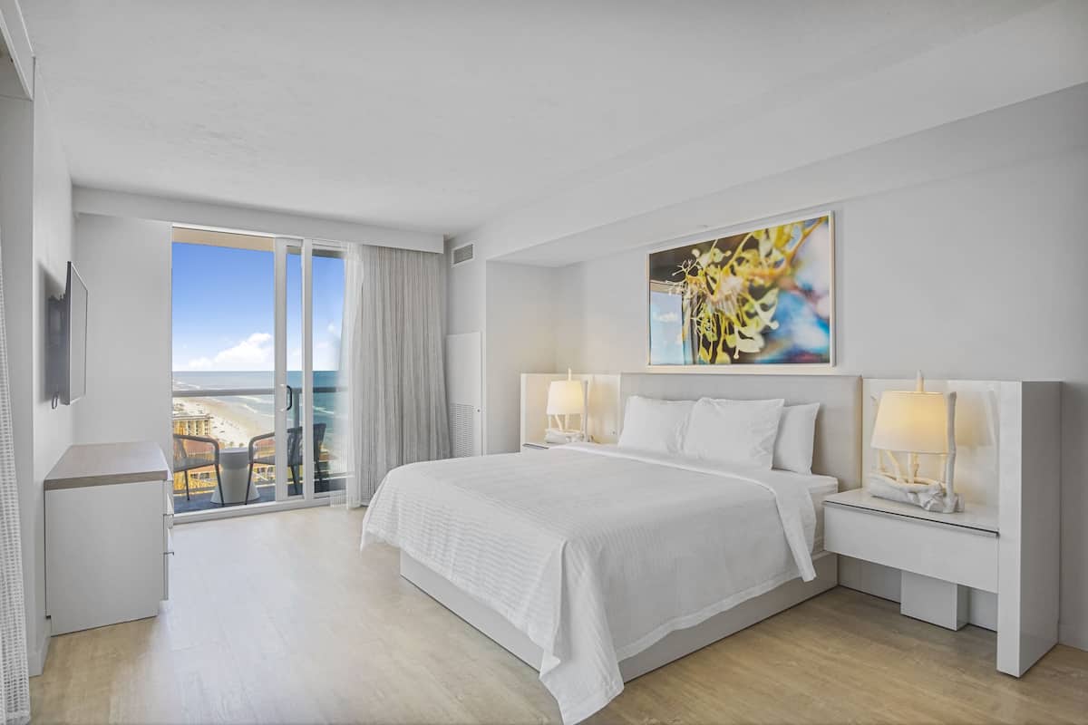 Ein Hotelzimmer mit einem großen Bett, zwei Nachttischen mit Lampen, einem Gemälde an der Wand und einem Balkon mit Blick auf den Strand.