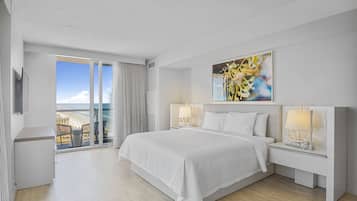 Quarto de hotel com uma cama grande, duas mesas de cabeceira com luminárias, um quadro na parede e uma varanda com vista para a praia.