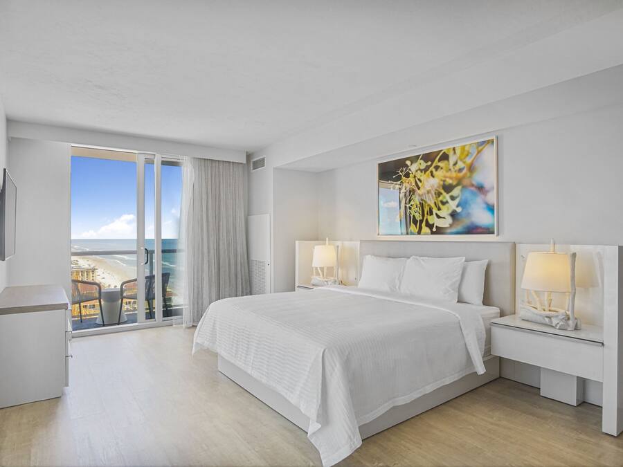 Ein Hotelzimmer mit einem großen Bett, zwei Nachttischen mit Lampen, einem Gemälde an der Wand und einem Balkon mit Blick auf den Strand.