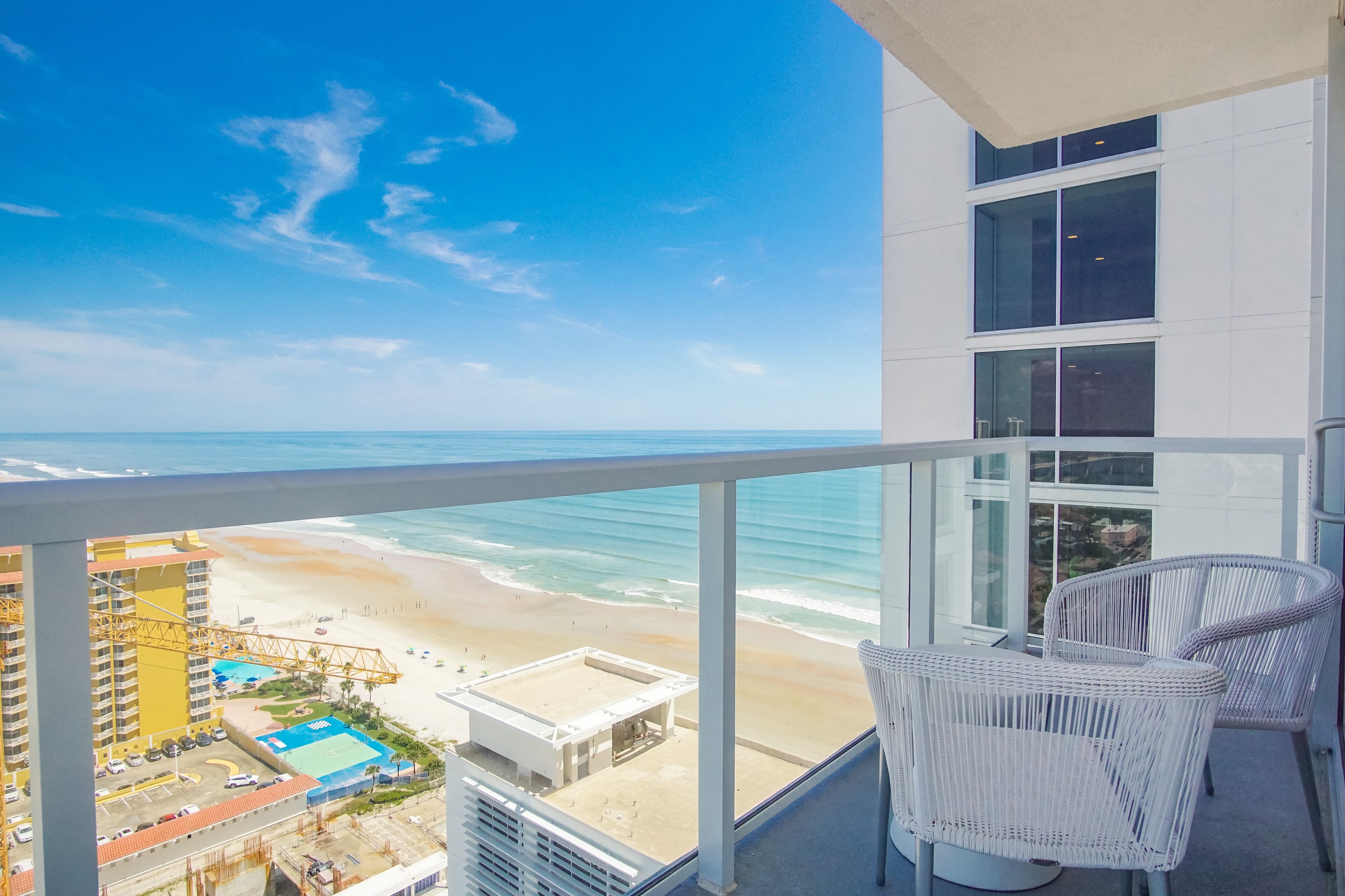 A balcony with a view of a beach and ocean. There are white wicker chairs and a glass railing.