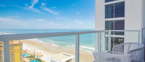A balcony with a view of a beach and ocean. There are white wicker chairs and a glass railing.