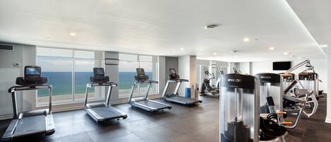A gym with treadmills, elliptical machines, and weight equipment, large windows with a view of the ocean.