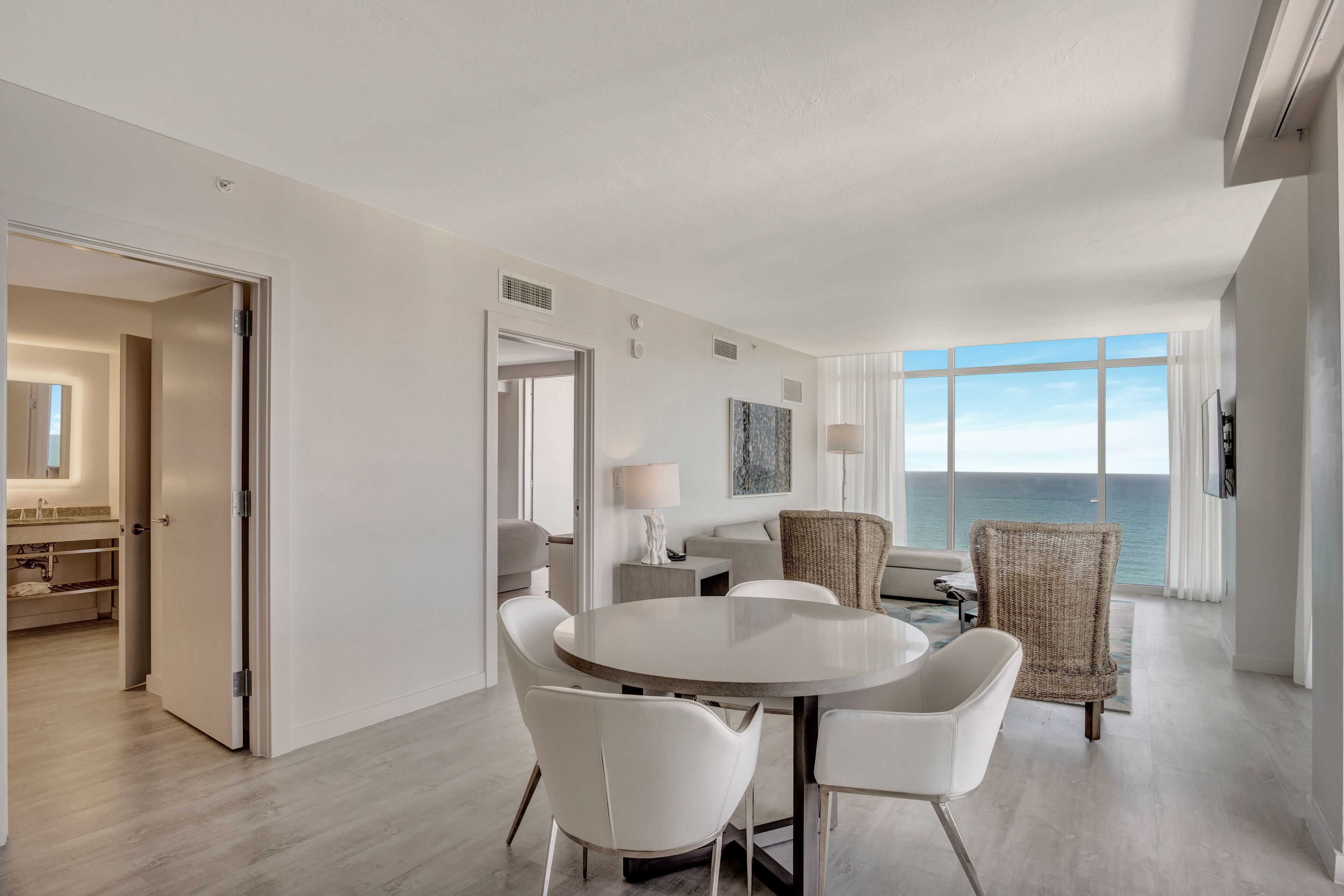 A modern hotel room with a round dining table, white chairs, a wicker armchair, and a view of the ocean through large windows.